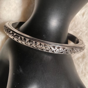 May include: A silver bangle bracelet with an ornate, scrolling design. The bracelet is round and has a detailed pattern around the entire circumference. The bracelet is displayed on a black mannequin hand.