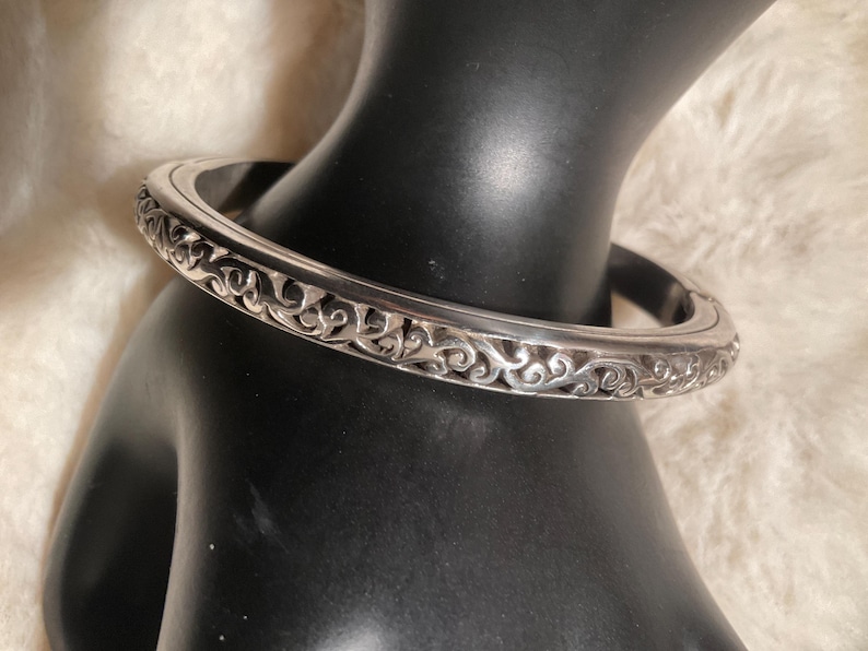 May include: A silver bangle bracelet with an ornate, scrolling design. The bracelet is round and has a detailed pattern around the entire circumference. The bracelet is displayed on a black mannequin hand.