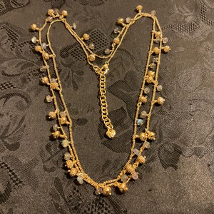 以下が含まれることがあります： 複数のストランドと小さな装飾ビーズが付いたゴールドトーンのネックレス。ネックレスは繊細なチェーンと調節可能な留め金が特徴です。ビーズはクリアとイリデッセントカラーのミックスで、微妙な輝きを加えています。ネックレスは暗い模様の表面に表示されています。