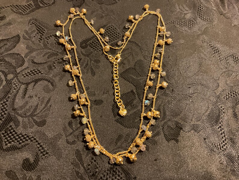 以下が含まれることがあります： 複数のストランドと小さな装飾ビーズが付いたゴールドトーンのネックレス。ネックレスは繊細なチェーンと調節可能な留め金が特徴です。ビーズはクリアとイリデッセントカラーのミックスで、微妙な輝きを加えています。ネックレスは暗い模様の表面に表示されています。