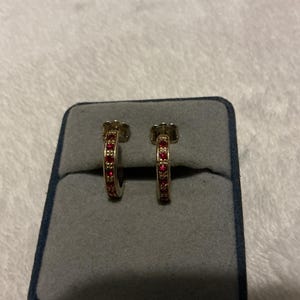 May include: A pair of gold hoop earrings featuring a row of small, vibrant red circular stones embedded along the front-facing surface. The earrings are displayed upright in a gray velvet-lined jewelry box with a dark blue outer frame.