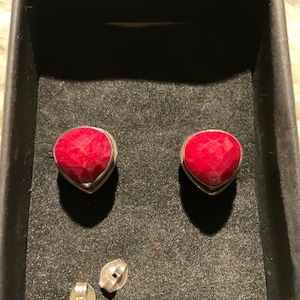 May include: A pair of heart-shaped ruby earrings with a silver setting, displayed in a black box. The earrings have a faceted surface, reflecting light. Two earring backs are also visible.