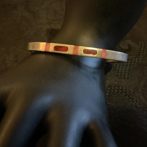 May include: Silver-toned bracelet with rectangular red and gold accents. The bracelet is displayed on a black mannequin hand, showcasing its design and color contrast. A simple, elegant piece of jewelry.