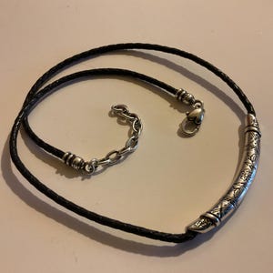 Puede incluir: Collar de cuero trenzado negro con un colgante curvo plateado. El colgante est&aacute; decorado con patrones grabados. El collar tiene una cadena plateada para ajustar la longitud y un cierre en forma de coraz&oacute;n.