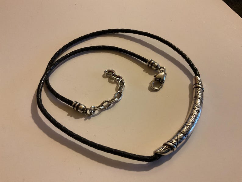 Puede incluir: Collar de cuero trenzado negro con un colgante curvo plateado. El colgante est&aacute; decorado con patrones grabados. El collar tiene una cadena plateada para ajustar la longitud y un cierre en forma de coraz&oacute;n.