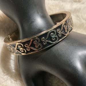 May include: A gold-coloured hinged bangle bracelet with a black enamel inlay. The bracelet features an ornate, swirling design in a contrasting gold colour. The jewellery is displayed on a black mannequin hand.