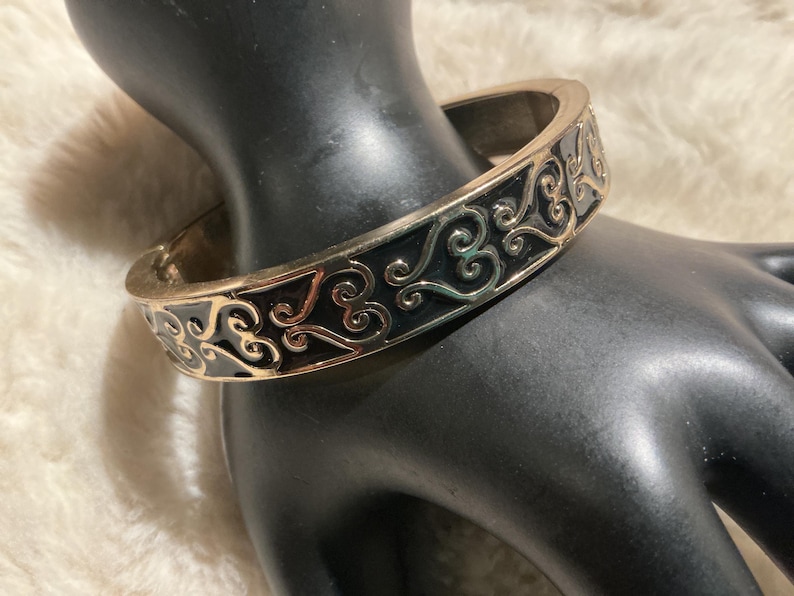 May include: A gold-coloured hinged bangle bracelet with a black enamel inlay. The bracelet features an ornate, swirling design in a contrasting gold colour. The jewellery is displayed on a black mannequin hand.