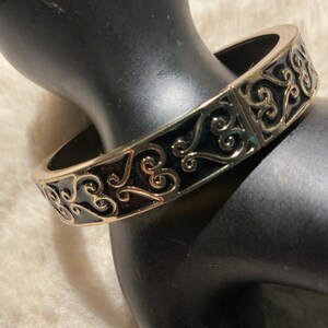 May include: A gold-coloured bangle bracelet featuring an elaborate, swirling filigree pattern. The bracelet has a black enamel inlay and a hinged clasp. The jewellery is displayed on a black mannequin hand.