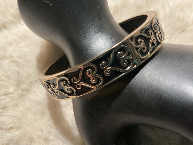 May include: A gold-coloured bangle bracelet featuring an elaborate, swirling filigree pattern. The bracelet has a black enamel inlay and a hinged clasp. The jewellery is displayed on a black mannequin hand.