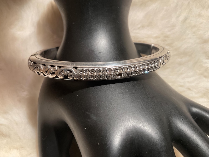 May include: Silver-coloured hinged bangle bracelet. The bracelet has two rows of small, clear crystals and decorative scrollwork. The jewellery is displayed on a black mannequin hand.