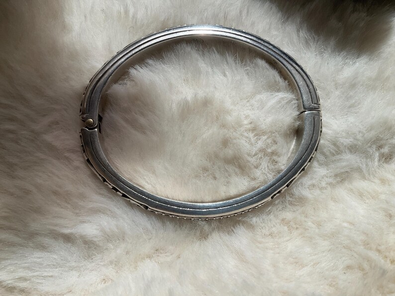 May include: Silver-coloured hinged bangle bracelet. The bracelet has a detailed design with small, dark accents along the edges. The bracelet is circular and appears to be made of metal.