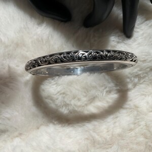 May include: Silver bangle bracelet featuring an intricate, swirling pattern. The bracelet is displayed on a white, fluffy surface. The dark design contrasts with the silver metal, creating a striking visual.