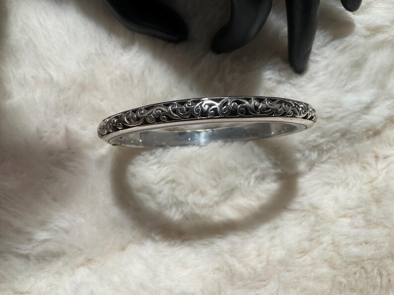 May include: Silver bangle bracelet featuring an intricate, swirling pattern. The bracelet is displayed on a white, fluffy surface. The dark design contrasts with the silver metal, creating a striking visual.