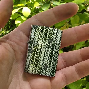 May include: A small, rectangular lighter with a decorative wave pattern in black and green, accented with black floral designs. The lighter is held in an open hand against a backdrop of green foliage.