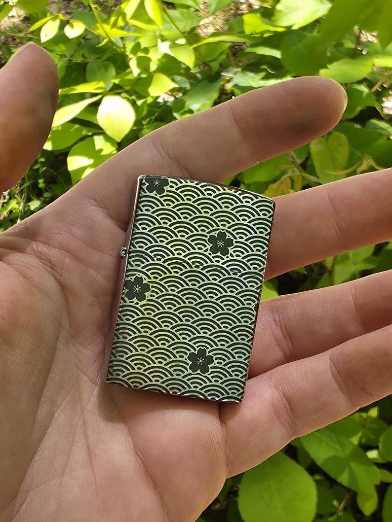 May include: A small, rectangular lighter with a decorative wave pattern in black and green, accented with black floral designs. The lighter is held in an open hand against a backdrop of green foliage.