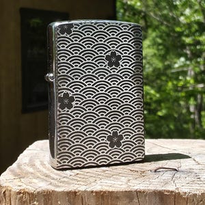 May include: A silver-colored lighter with a wave pattern and black flower accents. The lighter is rectangular and has a hinged lid. The design is etched into the metal, creating a contrast between the silver and black.