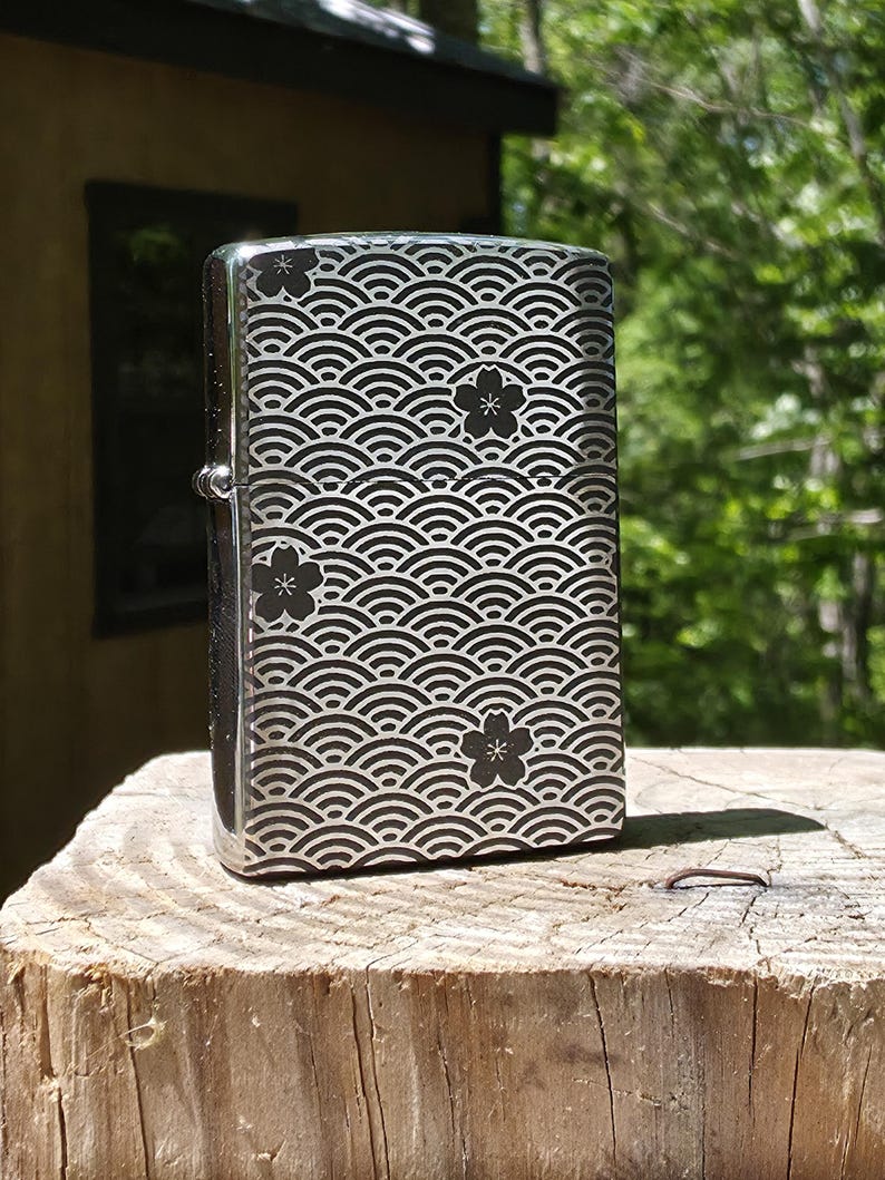 May include: A silver-colored lighter with a wave pattern and black flower accents. The lighter is rectangular and has a hinged lid. The design is etched into the metal, creating a contrast between the silver and black.