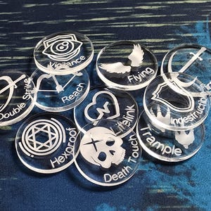 May include: A collection of clear acrylic game tokens with white etched designs and text. Tokens include "Vigilance", "Flying", "Double Strike", "Reach", "First Strike", "Hexproof", "Lifelink", "Death Touch", "Trample", and "Indestructible".