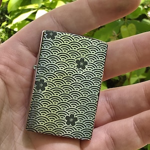 May include: A rectangular lighter with a wave pattern and black flower designs. The lighter has a metallic finish with a gradient of yellow and green. The design is repeated across the surface of the lighter.