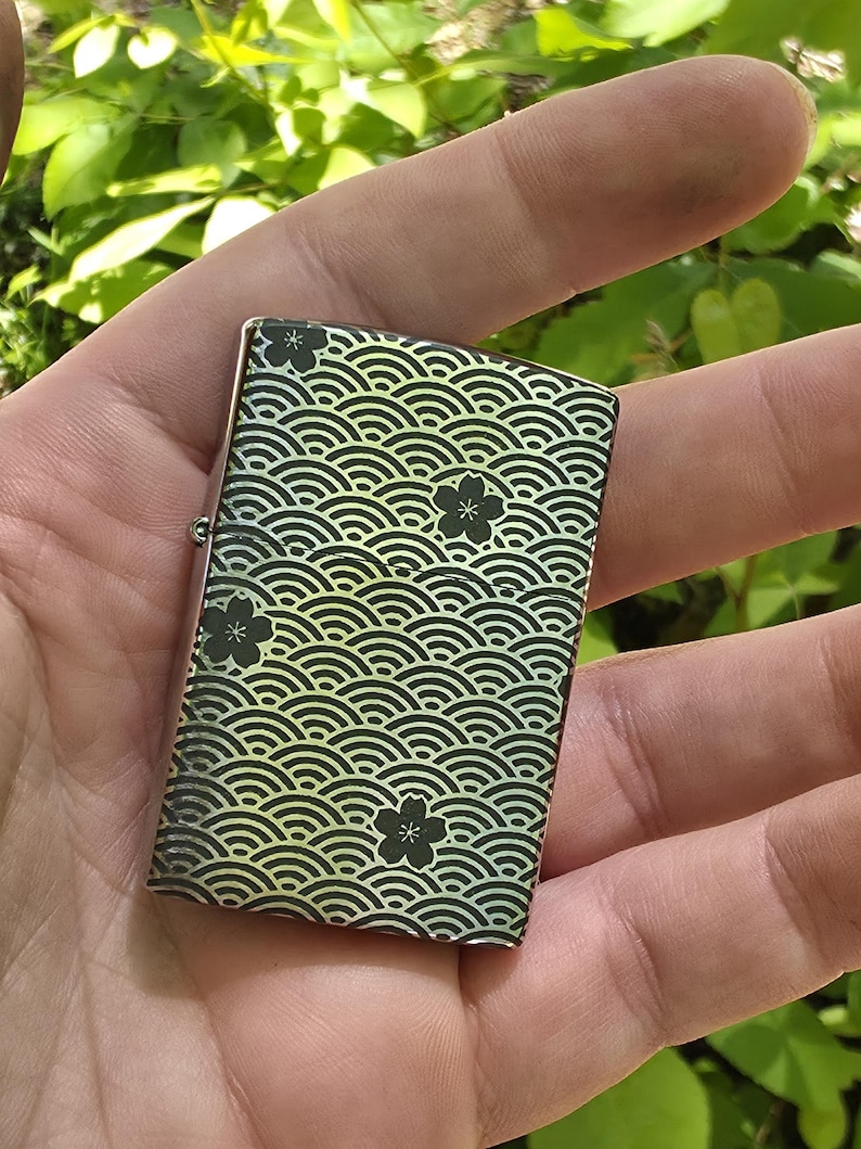 May include: A rectangular lighter with a wave pattern and black flower designs. The lighter has a metallic finish with a gradient of yellow and green. The design is repeated across the surface of the lighter.