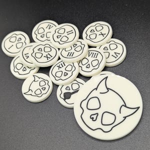May include: A collection of off-white, circular game tokens with black skull designs. Some tokens have Roman numerals. The tokens are scattered on a dark surface. The largest token has a detailed skull illustration.