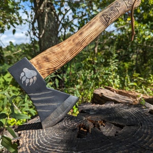 May include: A small, hand-forged axe with a black and white striped blade and a wooden handle. The handle is burned with a wood-burning design and the word "BEAR" is carved into the handle. The axe is resting on a tree stump.