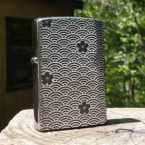 May include: A silver-colored lighter with a black wave pattern and floral accents. The design covers the entire surface of the lighter, creating a visually appealing and detailed aesthetic. The lighter is standing upright on a wooden surface.