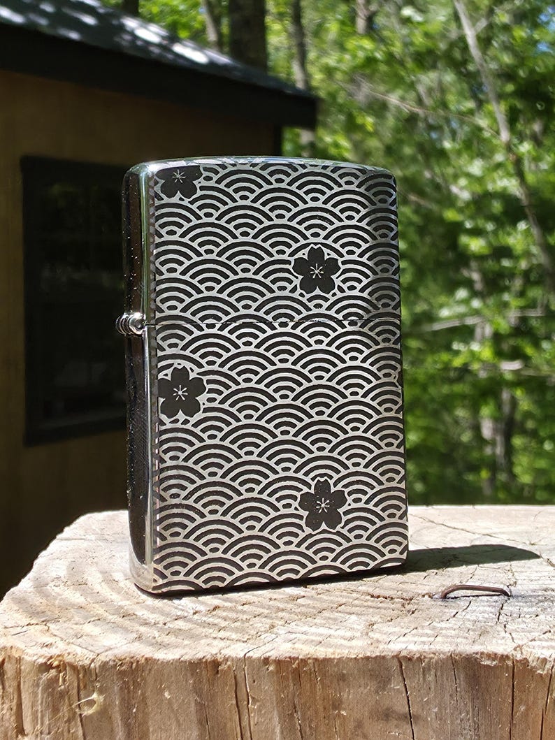 May include: A silver-colored lighter with a black wave pattern and floral accents. The design covers the entire surface of the lighter, creating a visually appealing and detailed aesthetic. The lighter is standing upright on a wooden surface.