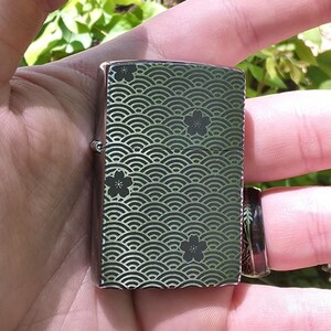 May include: A silver-colored lighter with a decorative pattern of curved lines and floral accents in black. The lighter is rectangular and has a hinged lid. The design covers the front and back of the lighter.
