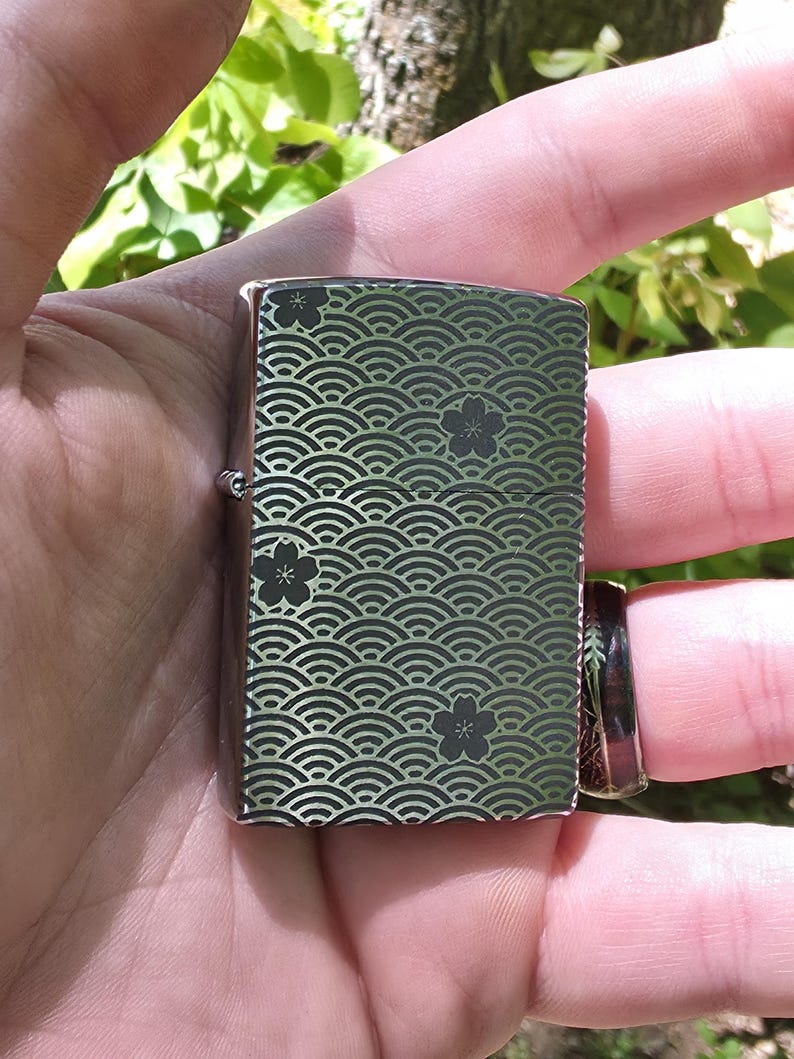 May include: A silver-colored lighter with a decorative pattern of curved lines and floral accents in black. The lighter is rectangular and has a hinged lid. The design covers the front and back of the lighter.