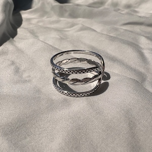 May include: A silver-colored ring with three bands. The top and bottom bands have a repeating diamond pattern, while the middle band features a twisted rope design. The ring is displayed on a light gray fabric.