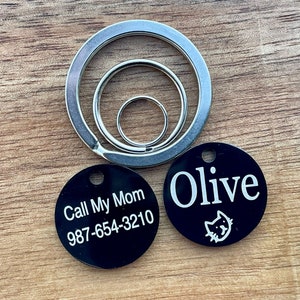 May include: Two black round pet ID tags with white text. One tag says "Call My Mom 987-654-3210" and the other tag says "Olive" with a small cat icon.