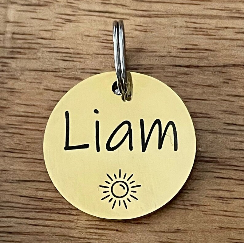 May include: Gold-colored round dog tag with a silver ring. The tag has the name "Liam" engraved on it and a small sun design below the name.