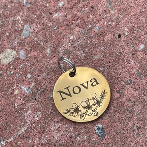 May include: Gold-colored round pet tag with the name "Nova" engraved in black lettering and a floral design.