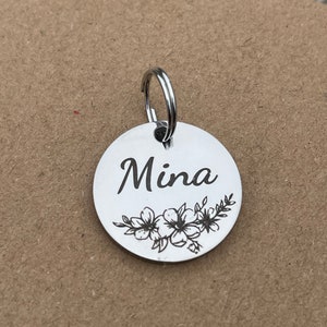 May include: Silver round tag with a floral design and the name "Mina" engraved on it. The tag has a silver ring attached to it.