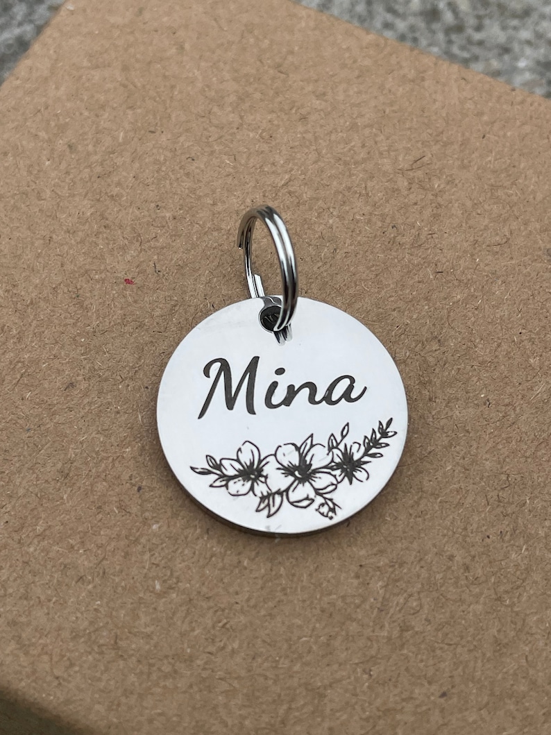 May include: Silver round tag with a floral design and the name "Mina" engraved on it. The tag has a silver ring attached to it.