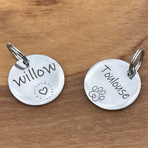 May include: Two silver dog tags with engraved names. The tag on the left says "Willow" with a heart and star design. The tag on the right says "Toulouse" with a paw print design.