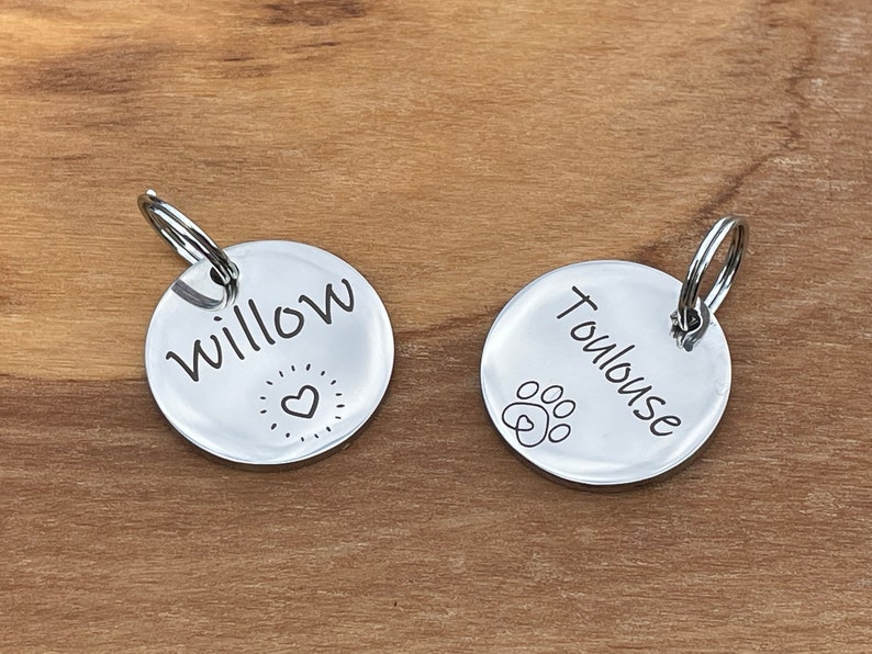 May include: Two silver dog tags with engraved names. The tag on the left says "Willow" with a heart and star design. The tag on the right says "Toulouse" with a paw print design.