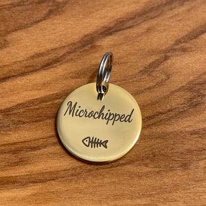 May include: A gold-colored round pet tag with a split ring. The tag has the text "Microchipped" engraved on it and a small fish bone symbol.