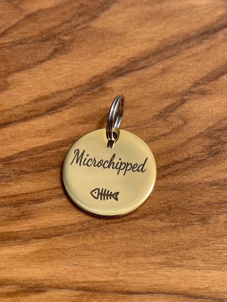 May include: A gold-colored round pet tag with a split ring. The tag has the text "Microchipped" engraved on it and a small fish bone symbol.