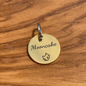 May include: A gold-colored round pet tag with the name "Mooncake" engraved on it. A small cat silhouette is engraved below the name.