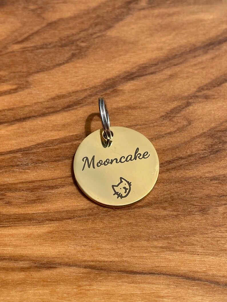 May include: A gold-colored round pet tag with the name "Mooncake" engraved on it. A small cat silhouette is engraved below the name.
