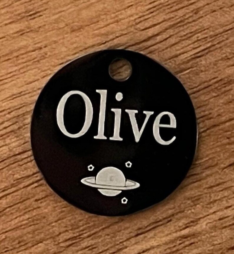 May include: Black round pet tag with the name "Olive" engraved in white lettering. A small white planet with rings and stars is engraved below the name.