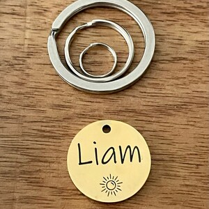 May include: Gold-colored round dog tag with the name "Liam" engraved on it and a small sun icon below the name. The tag is attached to a silver key ring with three interlocking circles.