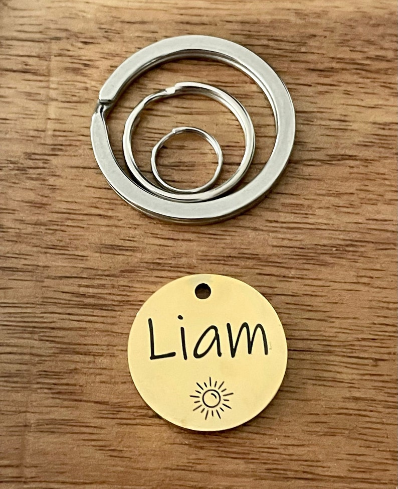 May include: Gold-colored round dog tag with the name "Liam" engraved on it and a small sun icon below the name. The tag is attached to a silver key ring with three interlocking circles.