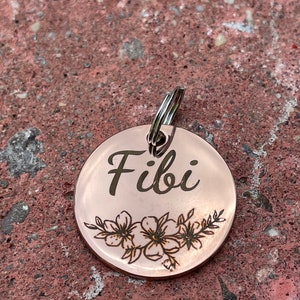 May include: A round, rose gold dog tag with the name "Fibi" engraved in a cursive font. The tag features a delicate floral design with small flowers and leaves.