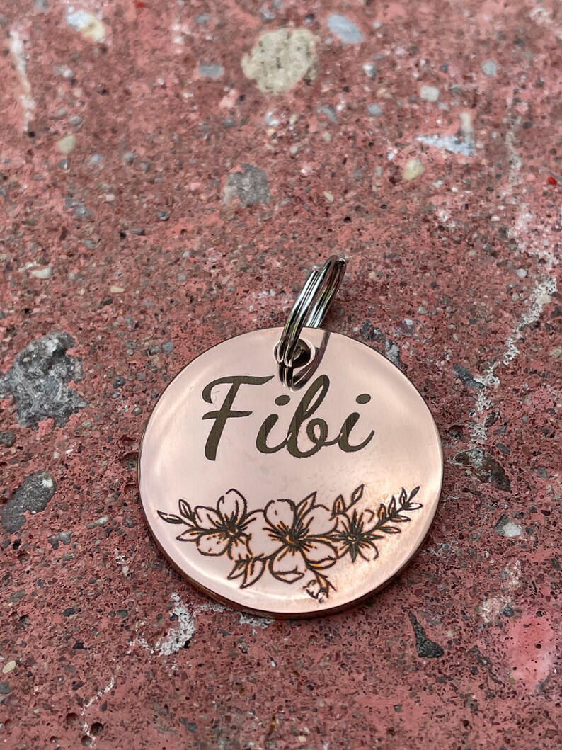 May include: A round, rose gold dog tag with the name "Fibi" engraved in a cursive font. The tag features a delicate floral design with small flowers and leaves.