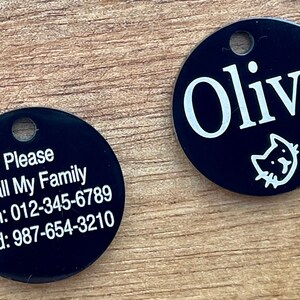 May include: Two black round pet ID tags with white text. One tag says "Please Call My Family Mom: 012-345-6789 Dad: 987-654-3210". The other tag says "Olive" with a small cat icon.