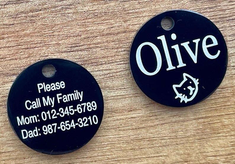 May include: Two black round pet ID tags with white text. One tag says "Please Call My Family Mom: 012-345-6789 Dad: 987-654-3210". The other tag says "Olive" with a small cat icon.