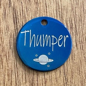 May include: Blue metal pet tag with the name "Thumper" engraved in white lettering. The tag also features a white silhouette of a planet with rings and stars.